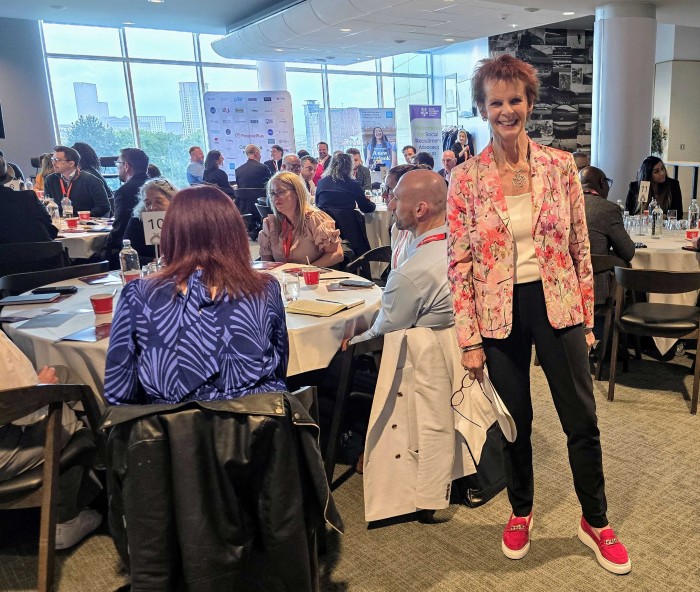 Rt. Hon. Anne Milton stands in a busy room of SRAG attendees at tables. She is smiling and projecting positivity that reflects the atmosphere on the day. 